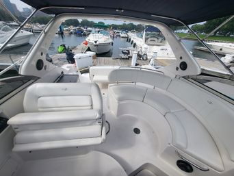Spacious white motor yacht cockpit with curved cushioned seating and fold-down helm seat under a canvas bimini, overlooking a busy dock with multiple boats at a tree-lined marina.