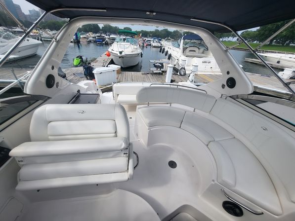Spacious white motor yacht cockpit with curved cushioned seating and fold-down helm seat under a canvas bimini, overlooking a busy dock with multiple boats at a tree-lined marina.