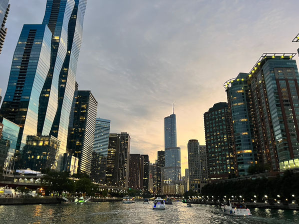 Sunset view of the Chicago River with boats cruising past illuminated downtown skyscrapers and reflective glass towers under a glowing sky