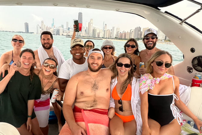 Group of friends laughing and posing on a boat in swimwear, with the Chicago skyline and lake in the background on a sunny summer outing.