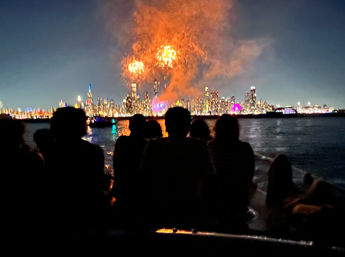 Silhouetted group on a boat watching bright orange fireworks burst over a glittering city skyline reflected on the water at night
