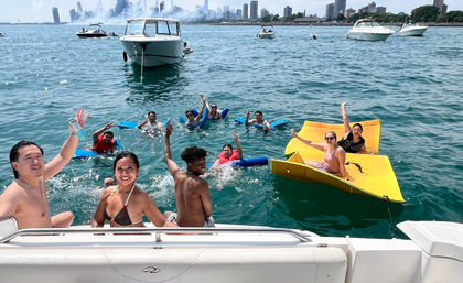 Group of people swimming and lounging on blue noodles and a bright yellow float near anchored boats on Lake Michigan with the Chicago skyline in the background, summer boating scene