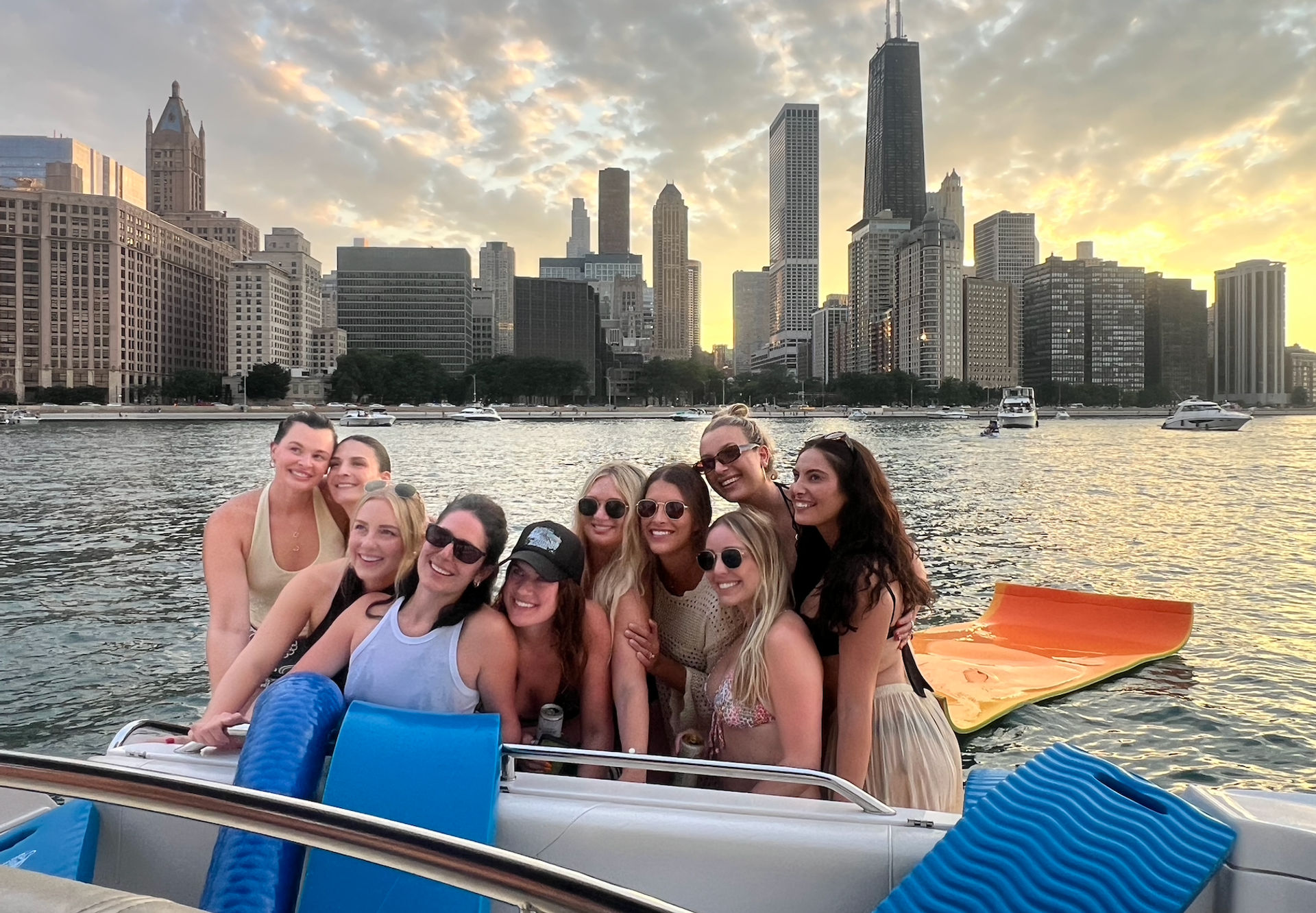 Group of friends smiling on a boat at sunset on Lake Michigan with the Chicago skyline and skyscrapers in the background, colorful inflatable float and yachts on the water