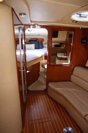 Cozy luxury yacht cabin interior with polished teak flooring, warm wood paneling, beige leather lounge, and a forward V-berth bed dressed with navy cushions and an anchor throw pillow.