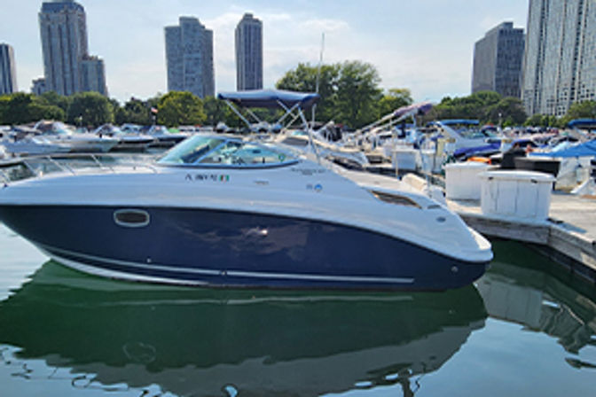 Sleek navy-and-white motorboat docked at an urban marina among other boats, with high-rise city skyline reflected in calm water on a sunny day.