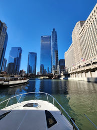 Bow of a white boat on the Chicago River with downtown glass skyscrapers and a historic riverside building under a bright blue sky.