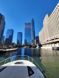 Bow of a boat on the Chicago River pointing toward downtown skyline with glass skyscrapers, a historic riverside building with flags, and bright blue sky reflected in the water.