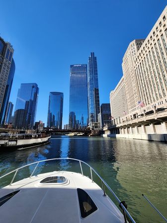 Bow of a boat on the Chicago River pointing toward downtown skyline with glass skyscrapers, a historic riverside building with flags, and bright blue sky reflected in the water.