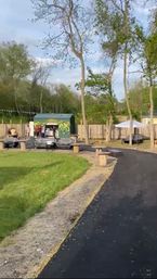 Tree-lined outdoor park with a green graffiti pop-up kiosk, people queuing, lounge sofas, wooden spool tables, bunting and a winding asphalt path beside a grassy lawn