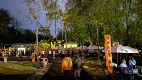 Evening outdoor night market in a wooded park — people stroll past tents, picnic tables and a colorful vertical wooden sign under string lights.