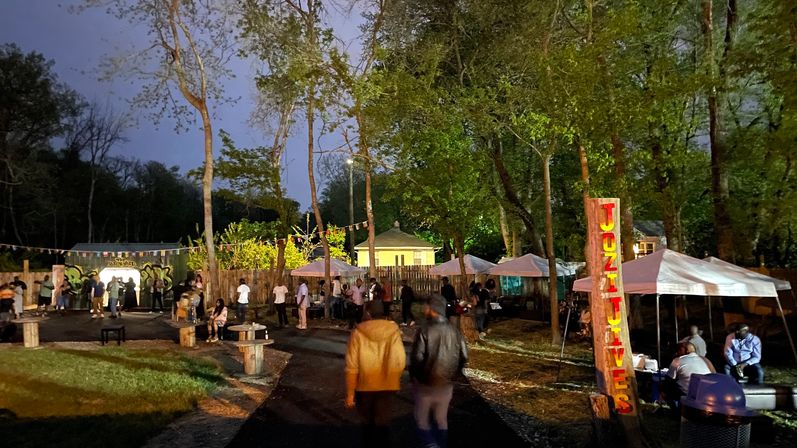 Evening outdoor night market in a wooded park — people stroll past tents, picnic tables and a colorful vertical wooden sign under string lights.