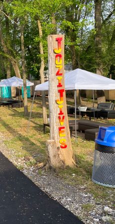 Bright red-and-yellow vertical letters mounted on a tree-trunk sign at a wooded outdoor event with white canopy tents, lounge seating, portable toilets, and a blue-topped trash can by an asphalt path.