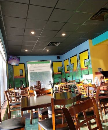 Bright, casual restaurant dining area with wooden tables and chairs, large front window, colorful mural lettering on blue and yellow walls, and black tiled ceiling with recessed lights