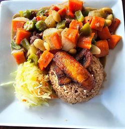 Caribbean-style plate of seasoned brown rice topped with fried plantain, chunky vegetable and bean stew with okra and carrots, and shredded cabbage slaw on a white square plate.