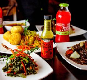 Jamaican-style Caribbean spread on white plates: scotch bonnet hot pepper sauce bottle, sautéed greens with peppers, fried dumplings, rich meat stew with root vegetables and a red tropical fruit drink.