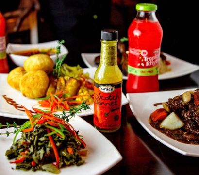 Jamaican-style Caribbean spread on white plates: scotch bonnet hot pepper sauce bottle, sautéed greens with peppers, fried dumplings, rich meat stew with root vegetables and a red tropical fruit drink.