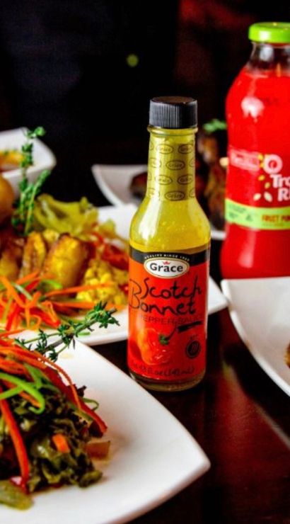 Jamaican-style Caribbean spread on white plates: scotch bonnet hot pepper sauce bottle, sautéed greens with peppers, fried dumplings, rich meat stew with root vegetables and a red tropical fruit drink.