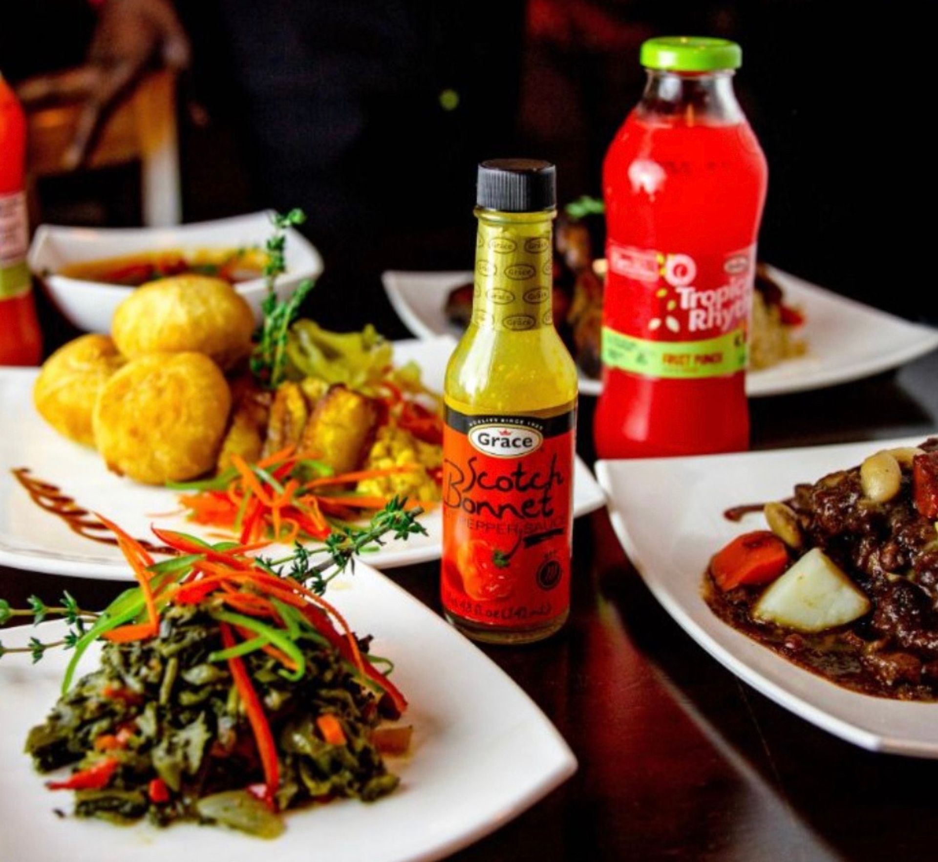 Jamaican-style Caribbean spread on white plates: scotch bonnet hot pepper sauce bottle, sautéed greens with peppers, fried dumplings, rich meat stew with root vegetables and a red tropical fruit drink.
