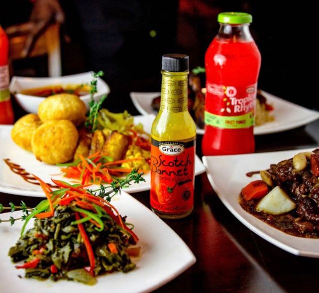 Jamaican-style Caribbean spread on white plates: scotch bonnet hot pepper sauce bottle, sautéed greens with peppers, fried dumplings, rich meat stew with root vegetables and a red tropical fruit drink.