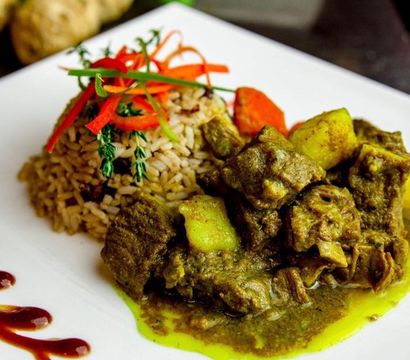 Hearty Jamaican curried goat with potatoes served beside seasoned rice, garnished with thyme and red pepper strips on a white plate