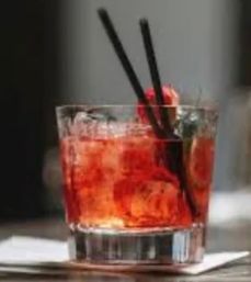 Close-up of a vibrant red cocktail in a short rocks glass with ice, two black stirrers and a cherry garnish resting on a napkin at a bar.
