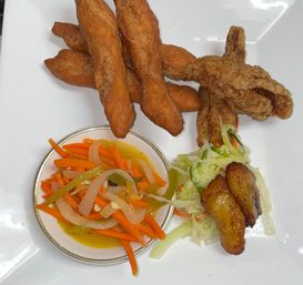 Caribbean-style plate with golden fried dough sticks, crispy fried chicken, pickled carrots and onions in a small bowl, shredded cabbage slaw and caramelized plantain slices on a white plate.