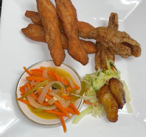 Caribbean-style plate with golden fried dough sticks, crispy fried chicken, pickled carrots and onions in a small bowl, shredded cabbage slaw and caramelized plantain slices on a white plate.