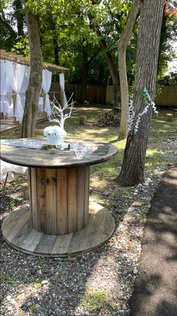 Charming backyard wedding scene with a rustic wooden spool table topped by a white floral centerpiece, draped pergola and surrounding trees.