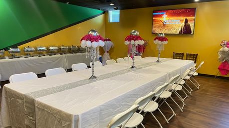 Indoor event space set for a party: long banquet table with white linens and silver sequin runner, pink floral and balloon centerpieces with crystal drops, white folding chairs, buffet chafing dishes along a yellow accent wall with wall-mounted TV.