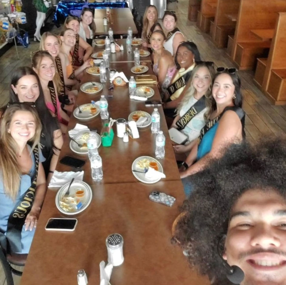 Smiling selfie of a sash-wearing group celebrating over brunch at a long wooden restaurant table with plates, bottled water and condiments.