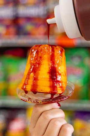Close-up of a Mexican-style mango paleta on a clear plastic holder, bright orange fruit ice being drizzled with red chamoy sauce from a squeeze bottle, held by a hand against blurred colorful store shelves.