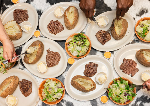 Overhead shot of a family-style meal with grilled steaks, baked potatoes topped with butter, and green side salads on plates over a cowhide tablecloth — hands reaching in to serve.