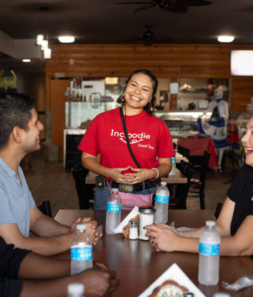 Smiling food tour guide chatting with a small group around a wooden table in a cozy restaurant, water bottles and condiments visible.