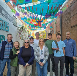 Eight people posing in a lively downtown alley beneath colorful papel picado banners and string lights, with brick walls and mural signage visible.