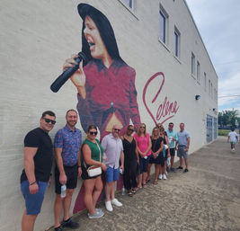 Group of people posing on a sidewalk in front of a large mural of a female singer holding a microphone with signature-style script on a white brick building.