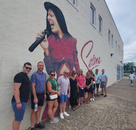 Group of people posing on a sidewalk in front of a large mural of a female singer holding a microphone with signature-style script on a white brick building.