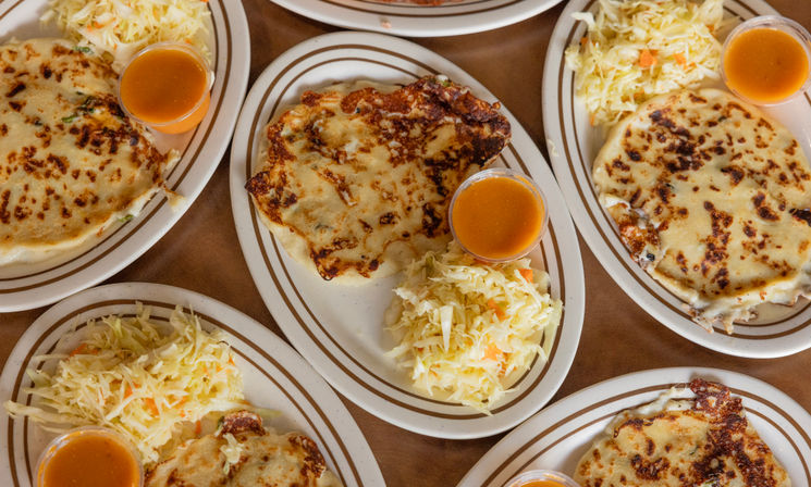 Top-down view of golden-grilled pupusas on oval plates served with shredded curtido coleslaw and bright orange salsa — Central American comfort food.