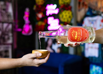 Hand pours clear spirit from a bottle with a red label into a small wooden cup in a colorful bar setting with neon sign and floral decor.