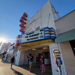 Retro neon movie theater marquee on a sunny downtown Texas main street, vertical TEXAS sign, people gathered under the entrance with blue sky and sun flare