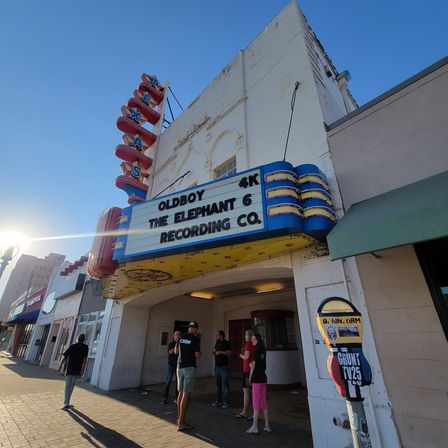 Retro neon movie theater marquee on a sunny downtown Texas main street, vertical TEXAS sign, people gathered under the entrance with blue sky and sun flare