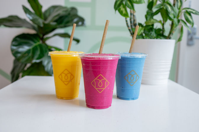 Vibrant yellow, magenta, and blue smoothies in clear takeaway cups with paper straws on a white countertop, potted plants and a light-green cafe interior in the background.