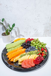 Vibrant fresh fruit platter on a black tray with sliced cantaloupe, honeydew, pineapple, grapefruit, kiwi, strawberries, blueberries, blackberries, raspberries and green grapes on a white tabletop with a small potted plant and textured white wall backdrop.