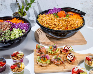 Vibrant catered spread: large bowl of seasoned noodles, colorful chopped salad with cucumbers and purple cabbage, assorted avocado and smoked-salmon crostini, and fruit-granola parfaits on a white table.