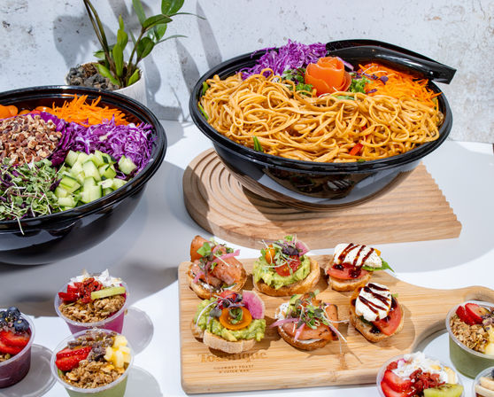 Vibrant catered spread: large bowl of seasoned noodles, colorful chopped salad with cucumbers and purple cabbage, assorted avocado and smoked-salmon crostini, and fruit-granola parfaits on a white table.