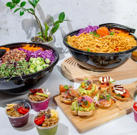Bright, healthy spread: large noodle bowl and vibrant salad bowl with purple cabbage, cucumber, nuts and microgreens, wooden board of avocado and smoked-salmon crostini, and small fruit granola cups on a white table.