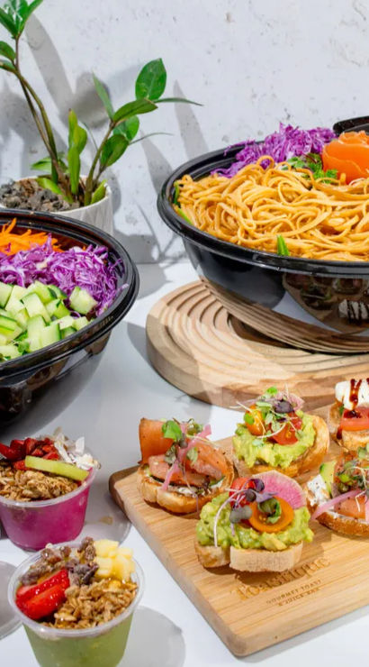 Bright, healthy spread: large noodle bowl and vibrant salad bowl with purple cabbage, cucumber, nuts and microgreens, wooden board of avocado and smoked-salmon crostini, and small fruit granola cups on a white table.