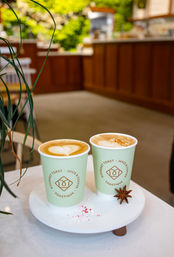Two lattes with heart latte art in mint takeaway cups on a white tray garnished with star anise and crushed peppermint, cozy cafe interior with wooden counter and greenery.
