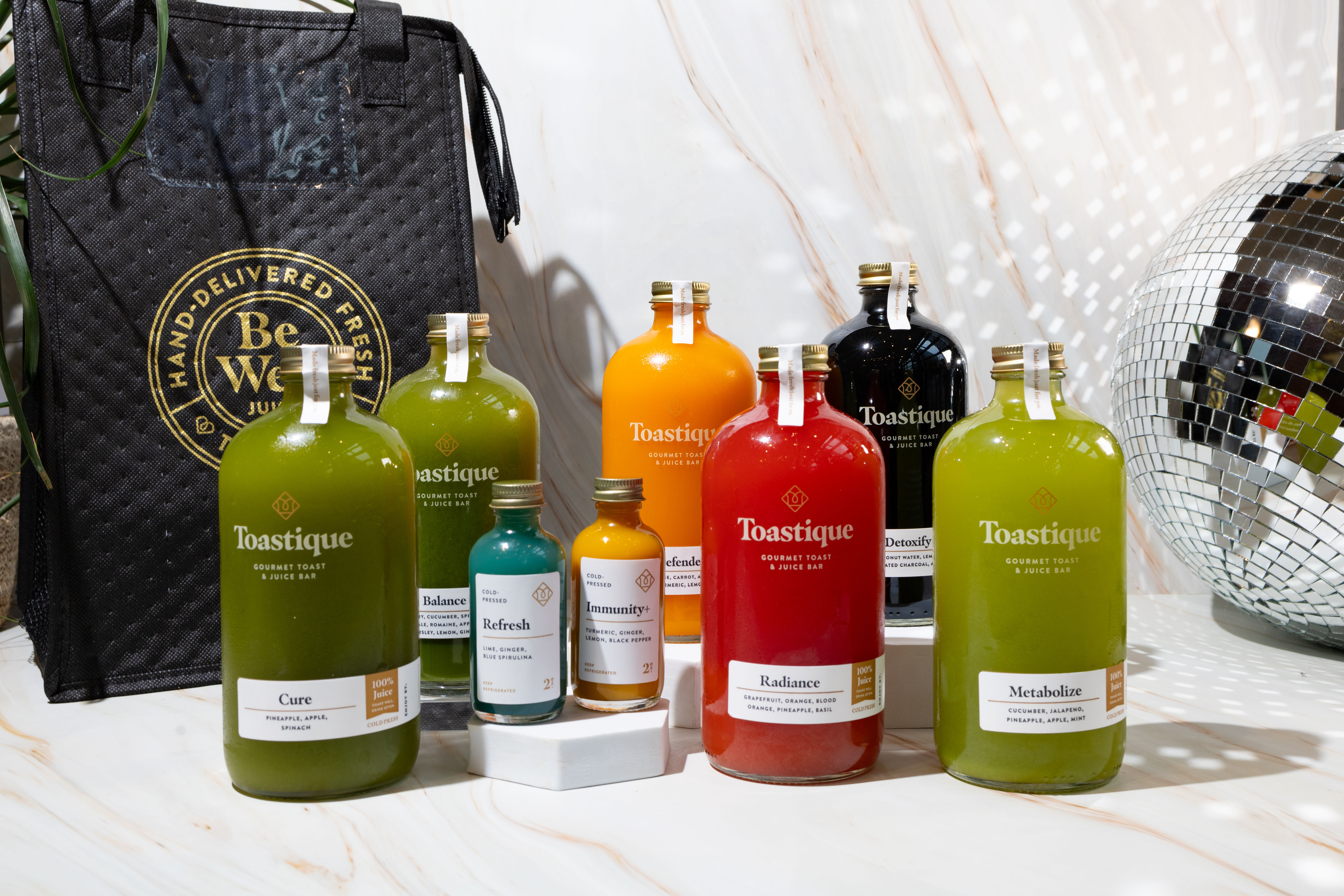 Vibrant glass bottles of cold-pressed juices and small wellness shots arranged on a marble countertop with a black insulated delivery bag and a mirrored disco ball in the background.