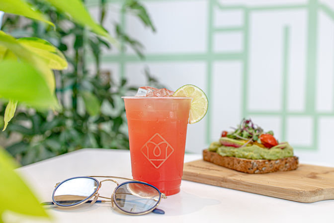 Refreshing pink iced drink with lime in a cup with a logo beside avocado toast topped with cherry tomatoes and microgreens on multigrain bread, blue-lensed sunglasses on a white table in a bright indoor café with green plants.