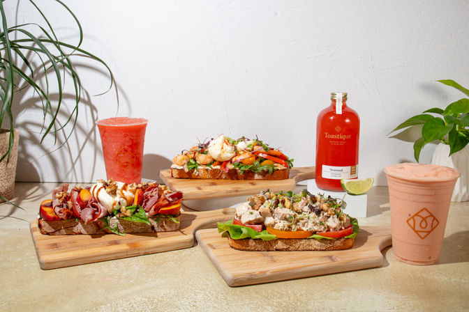 Assorted artisan open-faced toasts topped with burrata, shrimp, prosciutto, and chicken salad on wooden boards, paired with a strawberry smoothie and a cold-pressed red juice on a bright café countertop with potted plants.